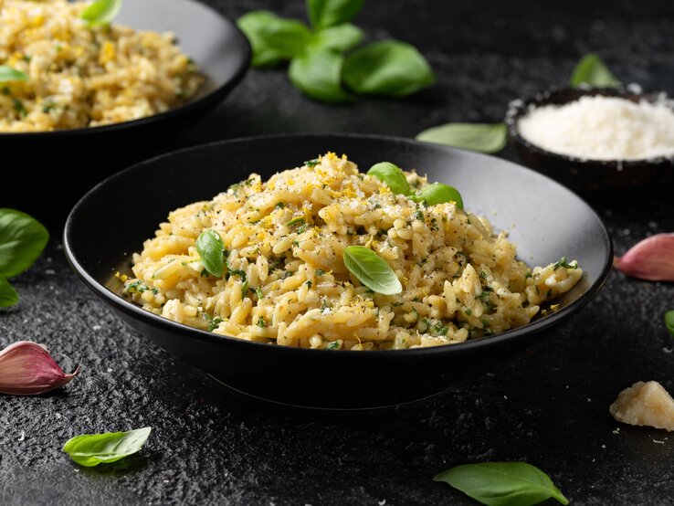 Frontalsicht: Auf einem schwarzen Untergrund stehen zwei dunkle runde Teller. In ihnen ist ein buttriges Zitronen-Orzo mit Parmesan. Drum herum liegen Basilikumblätter und Knoblauchzehen.