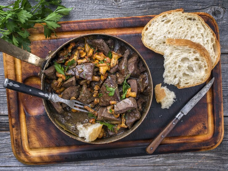 Low-Carb-Liebe: eine aromatische Leber mit Pfifferlingen. Draufsicht: auf dunklem Untergrund liegt ein Brettchen mit einer rustikalen Pfanne geratene Leber mit Pfifferlingen.