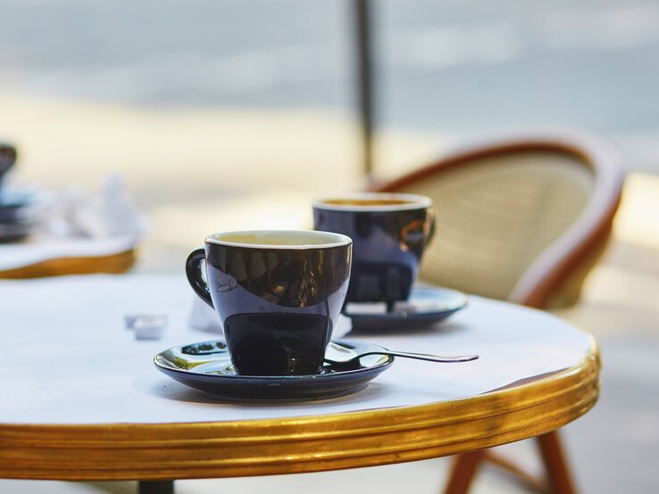 Frontalblick: Zwei French Hot Coffee in Tassen stehen auf einem runden Bistrotisch. An dem Tisch steht ein Stuhl.