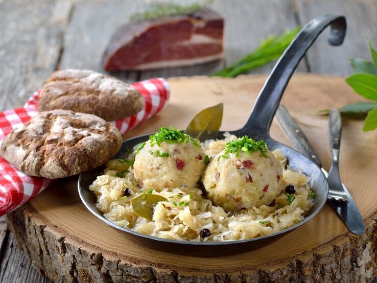 Purer Genuss: Sauerkraut mit selbst gemachten Speckknödeln versetzt zwar keine Berge – aber sie kommen näher. Ein Baumstamm steht auf einem Untergrund aus Holz. Darauf steht eine schmiedeeiserne Pfanne mit Sauerkraut und  selbst gemachten Speckknödeln. Daneben