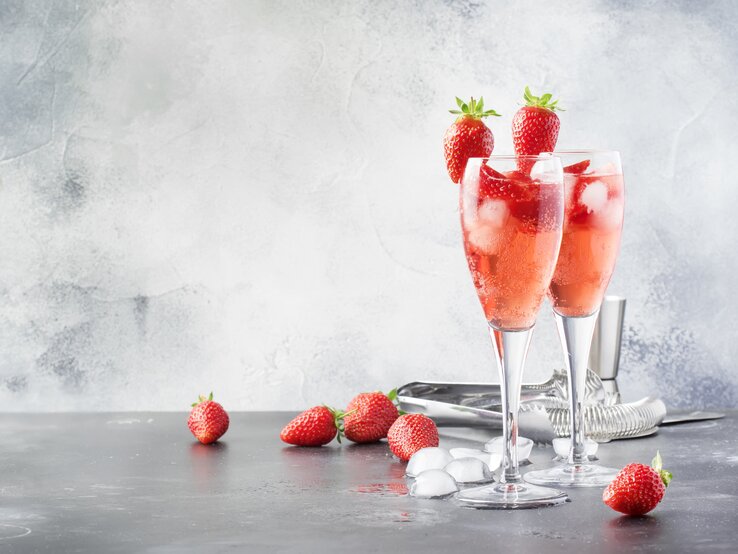 So schmeckt der Frühling besonders gut mit unserem Sekt mit Rhabarber und Erdbeeren! Zwei Gläser mit Rhabarber-Sekt und Erdbeeren stehen auf einem grauen Untergrund. Um die Gläser herum liegen Eiswürfel und Erdbeeren. Der Hintergrund ist mit einer Eisschicht überzogen. Hinter den Gläsern liegt eine Zange für die Eiswürfel.