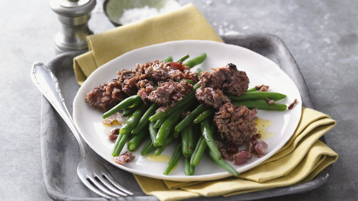 Ein weißer Teller mit der Hackfleischpfanne und en grünen Bohnen auf einem grauen Steintisch. Alles von schräg oben fotografiert.
