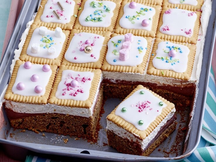 Blechkuchen mit Schokoladenboden, heller Creme und Butterkeksen mit weißem Zuckerguss und bunten Zuckerstreuseln, ein Stück ist herausgeschnitten.