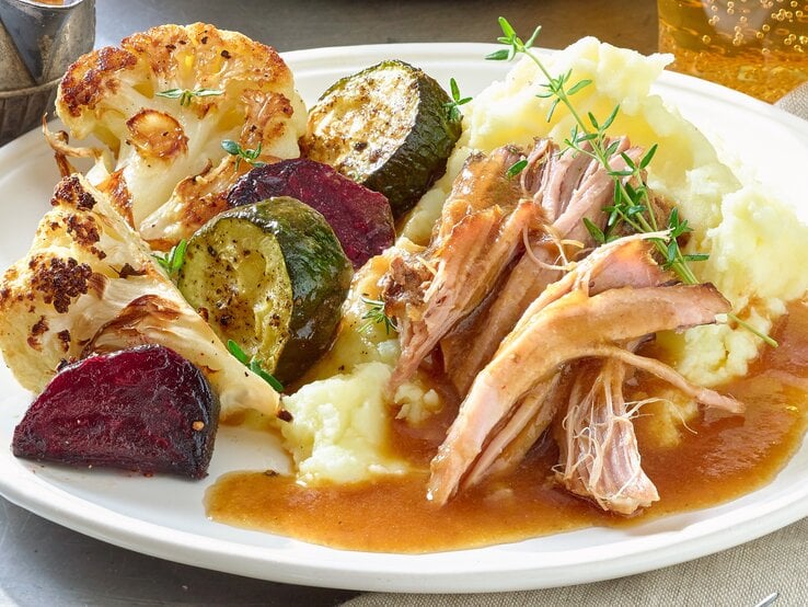 Pulled Pork Braten Pulled-Pork-Braten auf einem weißen Teller mit Kartoffelpüree, gebratenem Blumenkohl, Zucchini und roter Bete. Serviert mit Bratensoße. Der Teller steht auf einem Metalltisch neben einem grau-braunen Handtuch.