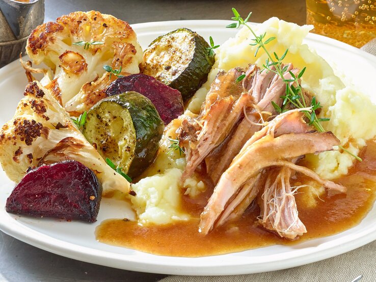Pulled Pork Braten Pulled-Pork-Braten auf einem weißen Teller mit Kartoffelpüree, gebratenem Blumenkohl, Zucchini und roter Bete. Serviert mit Bratensoße. Der Teller steht auf einem Metalltisch neben einem grau-braunen Handtuch.