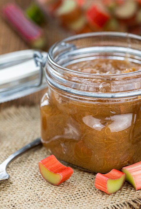 Ein Glas Rhabarber-Marmelade steht auf einem braunen Tuch auf einem Holztisch. Neben dem Glas liegt ein Löffel und vereinzelte Rhabarber-Stückchen.