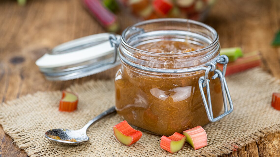 Ein Glas Rhabarber-Marmelade steht auf einem braunen Tuch auf einem Holztisch. Neben dem Glas liegt ein Löffel und vereinzelte Rhabarber-Stückchen.