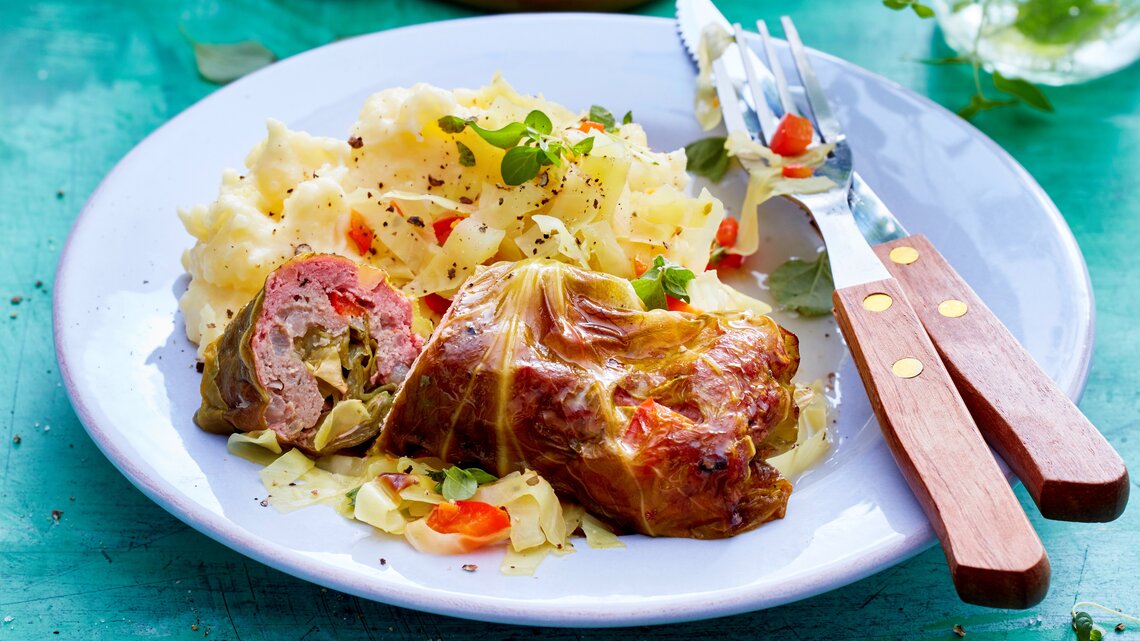 Spitzkohl-Rouladen an Kartoffelpüree auf einem weißen Teller und blauem Untergrund. Alles von schräg oben fotografiert.