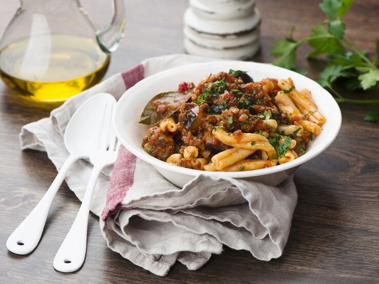 Nudeln-Lamm-Aubergine Ein weißer, tiefer Teller mit dem Pastagericht auf einem rustikalen Holztisch.
