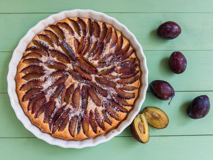 Eine weiße Kuchenform mit dem Pflaumenkuchen auf einem grünen Holztisch direkt von oben fotografiert.