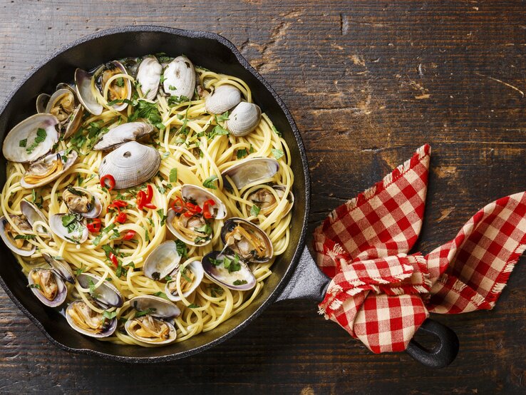 Spaghetti alle Vongole in einer gusseisernen Pfanne. Der Henkel der Pfanne ist mit einem rot-weiß karierten Küchentuch umwickelt. Das Ganze auf einem dunkelbraunen, hölzernen Untergrund.  | © StockFood / The Picture Pantry