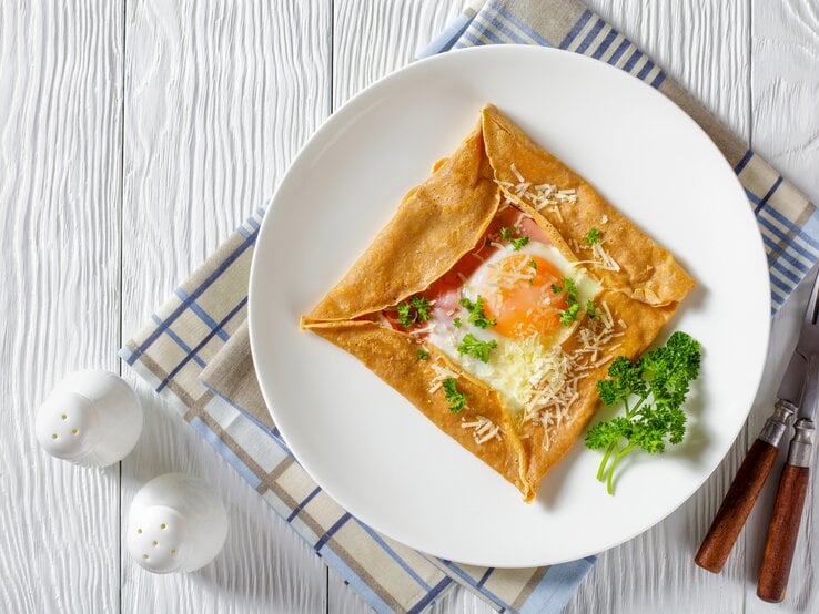 Aus der Vogelperspektive: Ein Teller mit einem Buchweizen-Pfannkuchen mit Schinken, Käse und Spiegelei  steht auf einem Küchentuch. Das Tuch liegt auf einem weißen Holztisch. Daneben stehen zwei Salzstreuer. Auch Messer und Gabel liegen dem dem Teller.