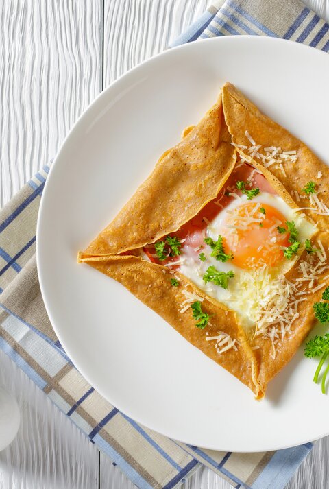Aus der Vogelperspektive: Ein Teller mit einem Buchweizen-Pfannkuchen mit Schinken, Käse und Spiegelei  steht auf einem Küchentuch. Das Tuch liegt auf einem weißen Holztisch. Daneben stehen zwei Salzstreuer. Auch Messer und Gabel liegen dem dem Teller.
