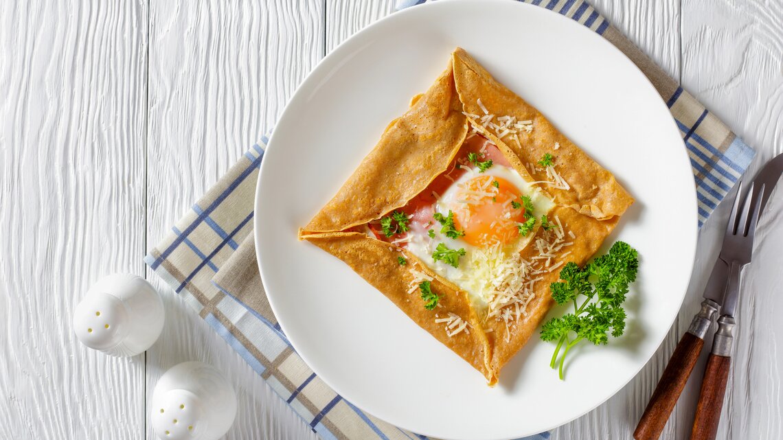Aus der Vogelperspektive: Ein Teller mit einem Buchweizen-Pfannkuchen mit Schinken, Käse und Spiegelei  steht auf einem Küchentuch. Das Tuch liegt auf einem weißen Holztisch. Daneben stehen zwei Salzstreuer. Auch Messer und Gabel liegen dem dem Teller.
