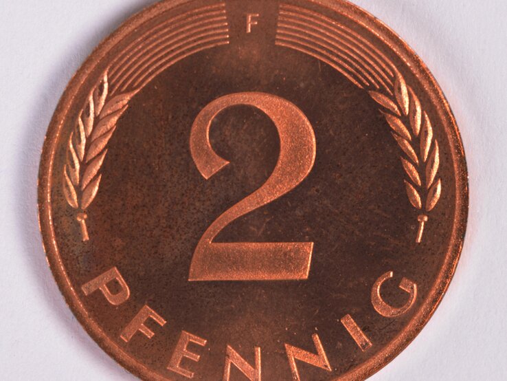 Das Bild zeigt eine 2-Pfennig-Münze aus Deutschland in Nahaufnahme. Die Münze hat eine kupferfarbene Oberfläche und ist zentral mit der Zahl "2" geprägt, die groß und deutlich zu sehen ist. Zu beiden Seiten der "2" befindet sich jeweils ein Ährenzweig. Oberhalb der Zahl befindet sich ein halbkreisförmiges Muster aus Strichen, und der Buchstabe "F" ist oben rechts zu erkennen, was auf die Prägestätte hinweist. Unten auf der Münze steht das Wort "PFENNIG". Der Hintergrund ist einheitlich weiß, was den Kontrast zur Münze erhöht und die Details hervorhebt. Die Münze zeigt leichte Gebrauchsspuren und Patina, was auf ihr Alter hindeutet. | © imago images / Schöning