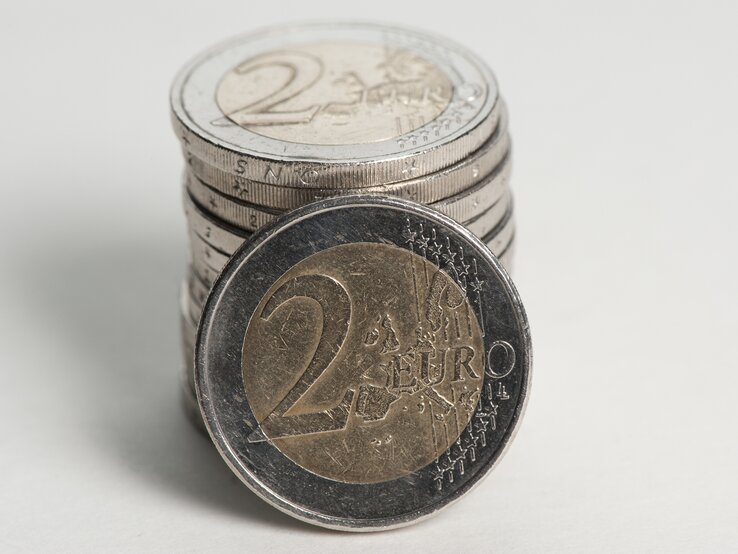 Das Bild zeigt eine Vorderansicht einer 2-Euro-Münze im Vordergrund, die auf ihrer Seite steht, mit einem Stapel weiterer 2-Euro-Münzen, die im Hintergrund unscharf zu sehen sind. Die Münze im Vordergrund hat einen silberfarbenen Kern und einen goldfarbenen Rand. Auf ihr ist das Nominal "2 EURO" zu erkennen, umgeben von einer geographischen Darstellung Europas und Sternen, die die Europäische Union symbolisieren.