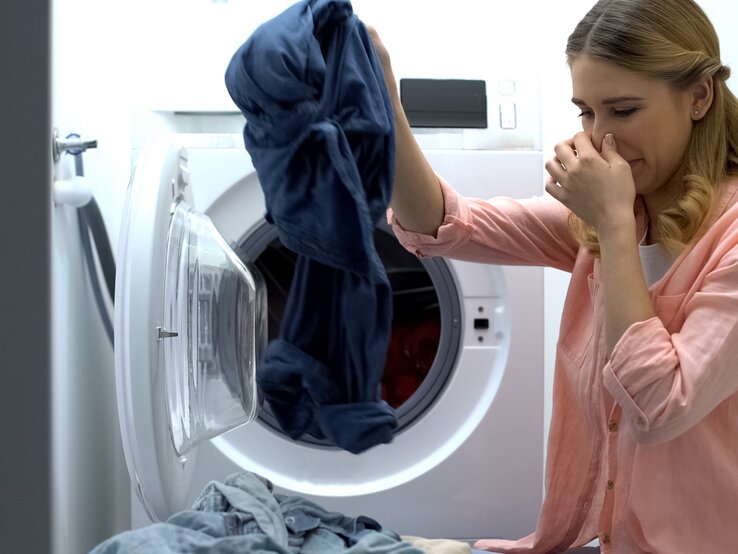 Frau holt mit der einen Hand Wäsche aus der Waschmaschine und hält sich mit der anderen die Nase zu