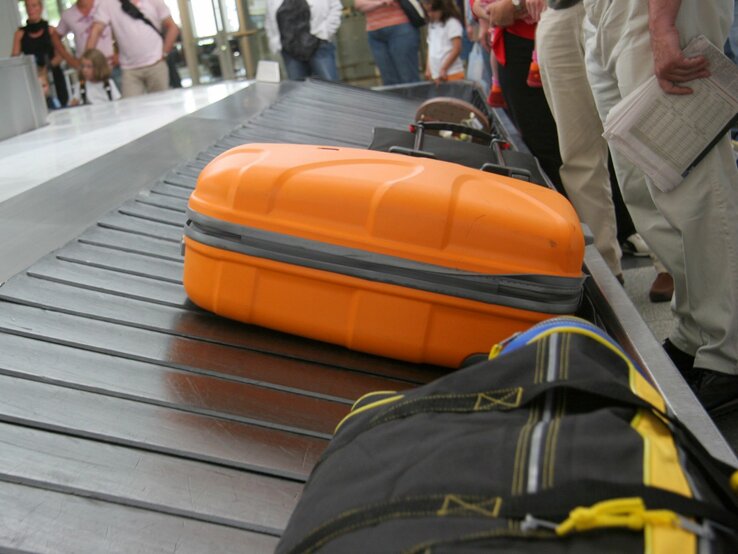  Das Bild zeigt einen Abschnitt eines Gepäckausgabebands an einem Flughafen aus einer niedrigen Perspektive. Im Vordergrund ist ein auffälliger orangefarbener Koffer zu sehen, der sich auf dem Band bewegt. Im Hintergrund sind unscharf weitere Gepäckstücke und wartende Passagiere zu erkennen. Die Menschen stehen oder gehen entlang des Bands, wahrscheinlich in Erwartung ihres eigenen Gepäcks. Der Fokus liegt auf dem orangefarbenen Koffer, was darauf hindeutet, dass die Aufnahme aus der Sicht eines wartenden Passagiers gemacht wurde.