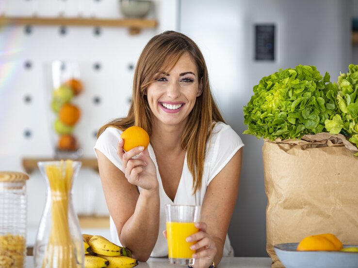 Das Bild zeigt eine fröhliche, junge Frau in einer modernen Küche, die eine Orange in der Hand hält und ein Glas Orangensaft auf dem Tisch vor sich hat. Sie hat langes, brünettes Haar und trägt ein weißes T-Shirt. Im Hintergrund sind eine Vase mit Spaghetti, eine Tüte mit frischem Salatkopf und eine Schüssel mit Zitrusfrüchten zu sehen. Die Frau lächelt in die Kamera, was die Assoziation von Gesundheit und Freude mit dem Verzehr von frischen Früchten und einer ausgewogenen Ernährung vermittelt.