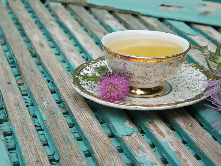 Eine Tasse Tee auf einer Holzunterlage, die Untertasse ist mit Pflanzen dekoriert | © iStock/TolikoffPhotography