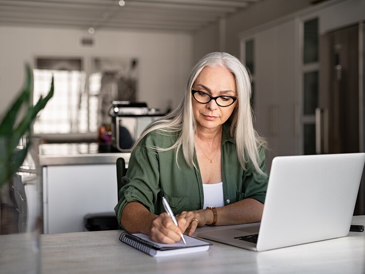 Eine konzentrierte ältere Frau mit langen grauen Haaren und Brille sitzt an einem hellen Arbeitsplatz und schreibt etwas in ein Notizbuch. Vor ihr steht ein geöffneter Laptop. Sie trägt ein olivgrünes Hemd und wirkt, als wäre sie gerade in einer Arbeits- oder Lernumgebung zu Hause. Das Zimmer ist hell und modern eingerichtet, mit einer Küche im Hintergrund, was auf ein häusliches Büro hindeutet. Das natürliche Licht und die Pflanze auf dem Tisch schaffen eine angenehme und ruhige Arbeitsatmosphäre. | © iStock.com/Ridofranz