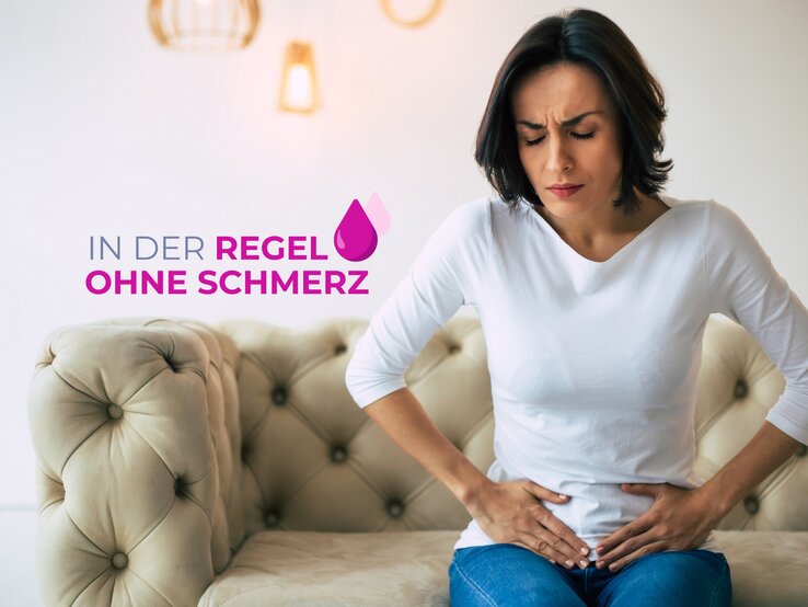 Eine Frau mittleren Alters mit dunklen Haaren sitzt auf einer cremefarbenen Couch und hält sich den Unterbauch, während sie schmerzverzerrt nach unten blickt. Sie trägt ein schlichtes weißes Oberteil und blaue Jeans. Ihr Gesichtsausdruck deutet auf Unbehagen und Bauchschmerzen hin. Über ihr schwebt der Text "IN DER REGEL OHNE SCHMERZ" in Großbuchstaben, begleitet von einem Tropfensymbol. Der Hintergrund ist hell und modern gestaltet, mit dezenten Wandleuchten, die eine warme und einladende Atmosphäre erzeugen.