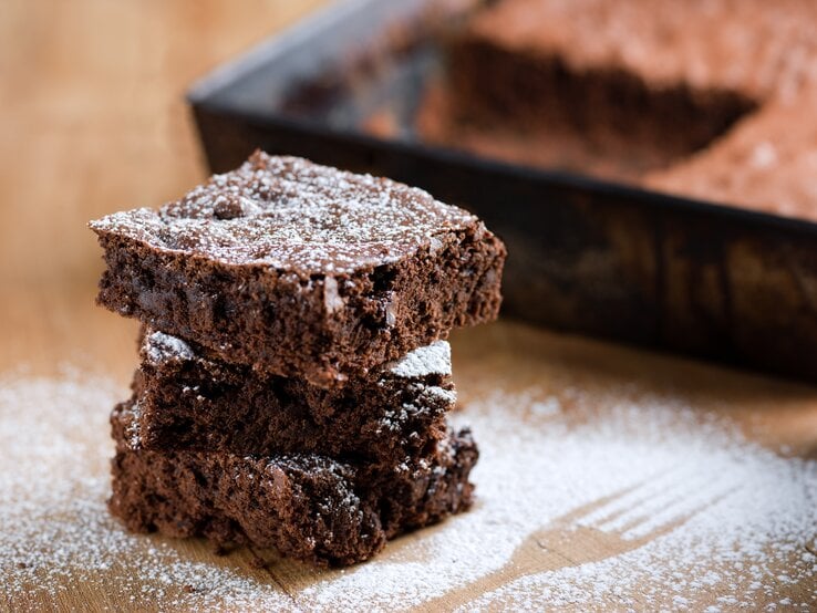 Mehrere angeschnittene Stücke Schokoladenkuchen mit Puderzucker bestreut auf Holztisch. 