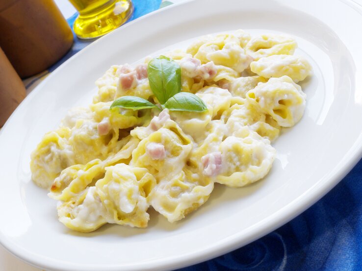 Auf dem Bild ist ein Teller mit Tortellini in einer cremigen Sauce zu sehen. Die Pasta ist mit kleinen Schinkenwürfeln angereichert und mit frischem Basilikum garniert. Der Teller steht auf einem Tisch mit einer blauen Tischdecke und im Hintergrund ist eine Flasche Olivenöl angedeutet. Das Gericht wirkt frisch und ist klassisch italienisch angerichtet.