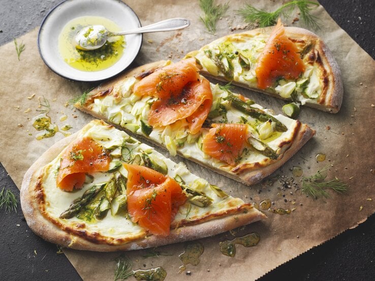 Flammkuchen mit grünem Spargel und Räucherlachs auf Backpapier und dunklem Tisch.