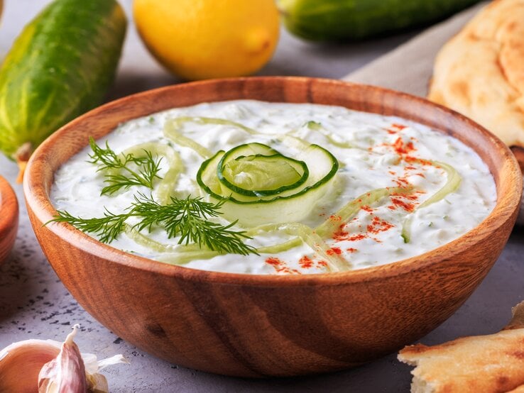Frischer Zaziki mit Gurke in einer Holzschale. Daneben liegen Gurken, Knoblauch, Zitronen und Brot. 
