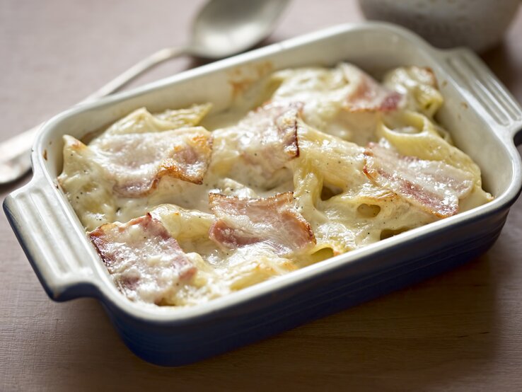 Nudelgratin Cremiger Nudelauflauf mit geschmolzenem Käse und knusprigem Schinken in einer blauen Keramikform auf einem Holztisch.