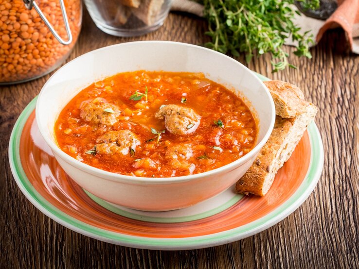 Linsen-Tomatensuppe mit Hackbällchen / runde Schüssel auf Unterteller / zwei Brotscheiben / Weckglas mit roten Linsen / Frische Kräuter / Untergrund Holz / Draufsicht, schräg