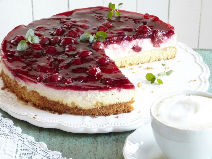 Die angeschnittene Kirsch-Schmand-Torte seitlich auf einem weißen Kuchenteller und hellem Tisch.