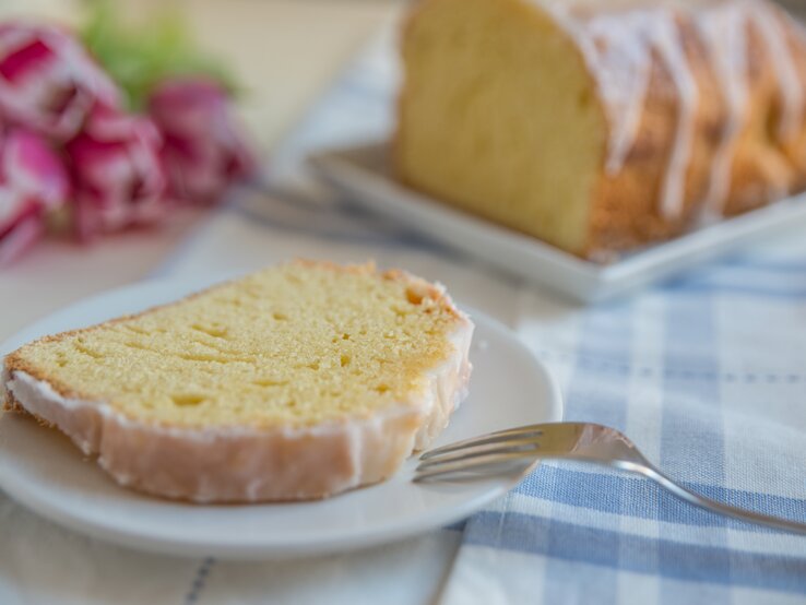 Ein Stück vom Zitronenkuchen auf einem weißen Teller mit hellblau-weiß-karierter Tischdecke. Alles nah und von der Seite fotografiert.