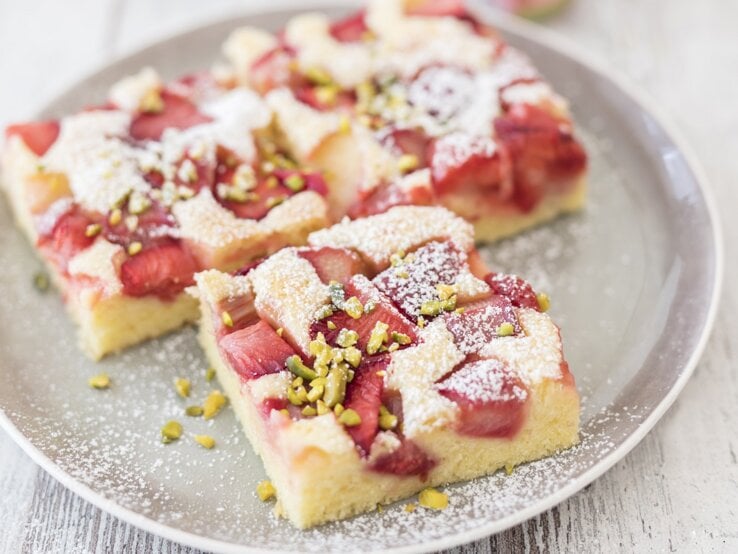 Für alle Rhabarberfans: Unser schnelles und einfaches Rezept für köstlichen Rhabarberkuchen vom Blech. Drei quadratische Stücke Rhabarberkuchen vom Blech auf einem runden Teller.
