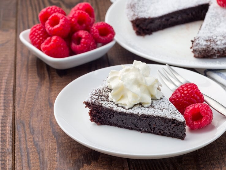Schokoladenkuchen mit Puderzucker, Sahne und Himbeeren auf weißem Teller in rustikalem Ambiente. 