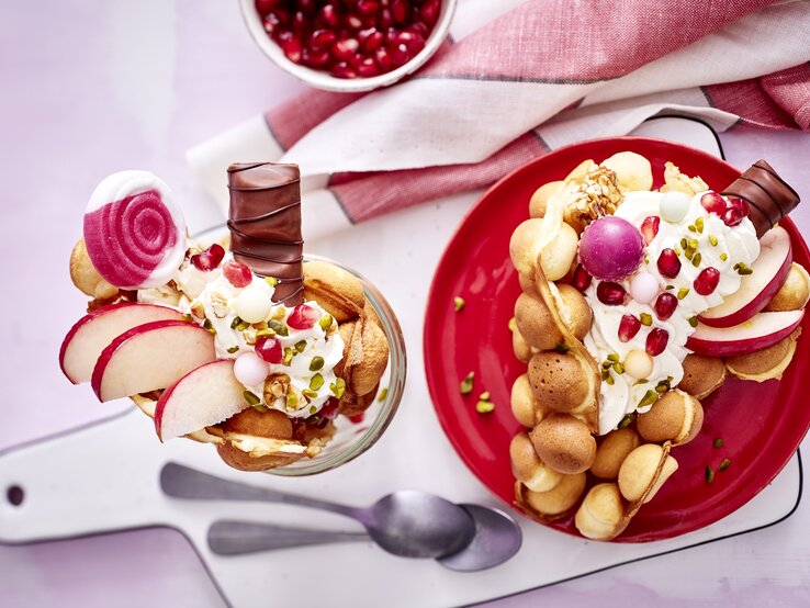 Eine große Bubble-Waffel auf einem runden Teller / eine kleine im Glas / zwei Teelöffel / Brettchen / Küchentuch / Draufsicht