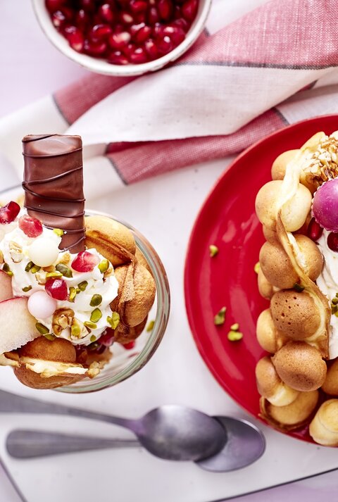 Eine große Bubble-Waffel auf einem runden Teller / eine kleine im Glas / zwei Teelöffel / Brettchen / Küchentuch / Draufsicht