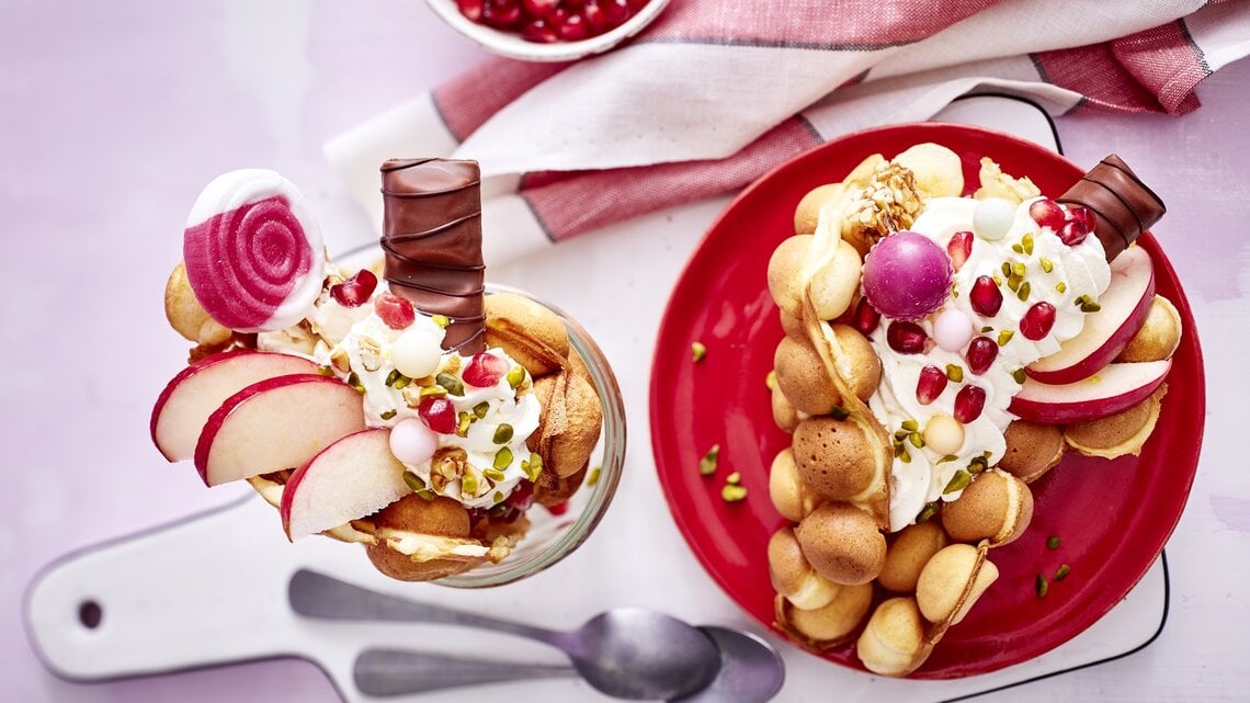 Eine große Bubble-Waffel auf einem runden Teller / eine kleine im Glas / zwei Teelöffel / Brettchen / Küchentuch / Draufsicht