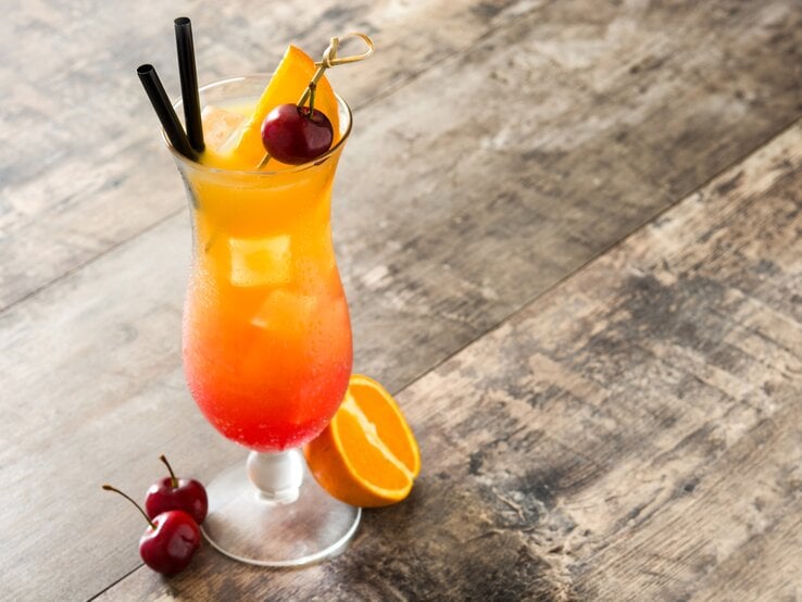 Liebevoll dekoriertes Glas gefüllt mit einem fruchtig aussehendem Cocktail auf Holztisch. 