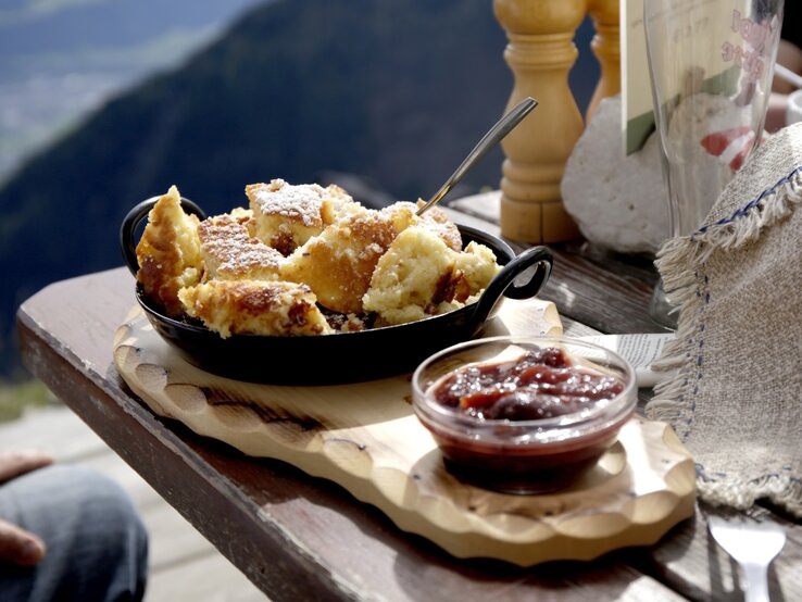 Bananenschmarrn in Pfanne auf Holzbrett, daneben Kompott Schälchen