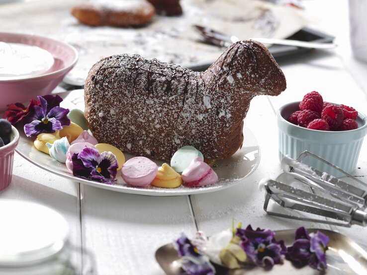 Auf einem hellen Holztisch liegt ein gebackenes Osterlamm mit Baisertupfen.
