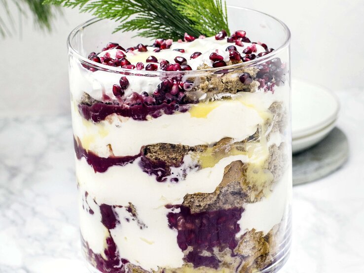 Das Lebkuchen-Trifle mit Eierlikör im Glas vor hellem Untergund mit weihnachtlicher Deko garniert. Alles sehr nah und direkt von vorne fotografiert.
