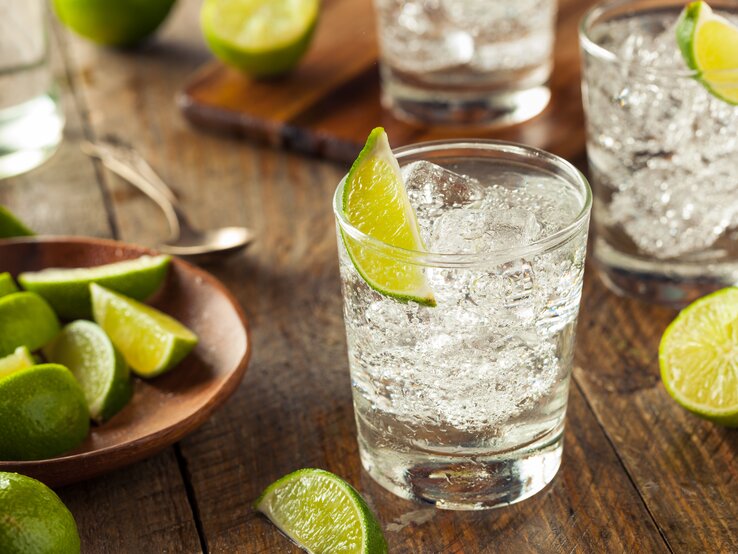 Gin Tonic Glas mit prickelnder, durchsichtiger Flüssigkeit (Gin Tonic) dekortiert mit Limette auf Holztisch