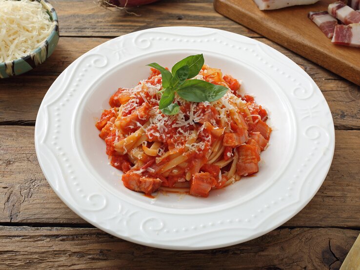 Teller mit Pasta in Tomatensoße, bestreut mit geriebenem Käse und gekrönt mit einem frischen Basilikumblatt, serviert auf einem rustikalen Holztisch. 