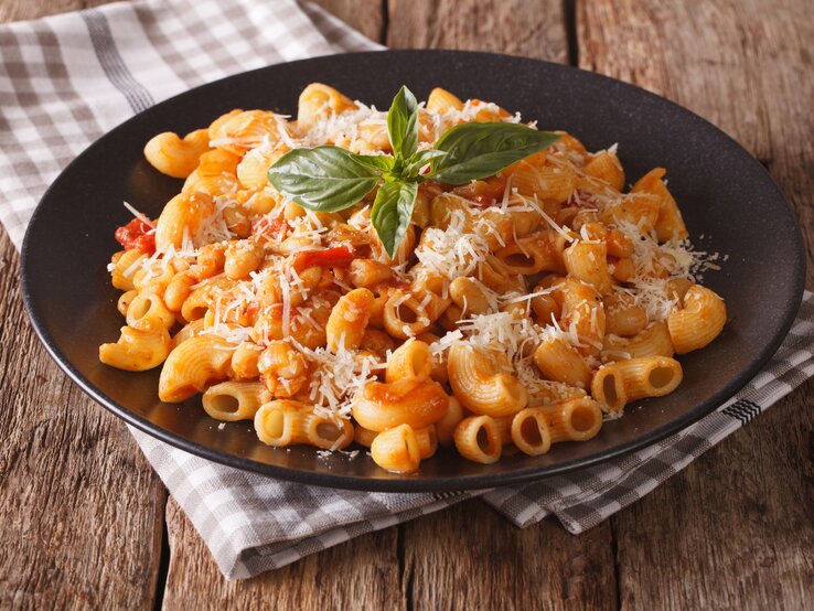 kurze Röhrennudeln in einer Bohnen-Tomaten-Soße auf schwarzem Teller, bestreut mit geriebenem Käse. Das Ganze steht auf einem rustikalem Holztisch.  | © iStock/ ALLEKO