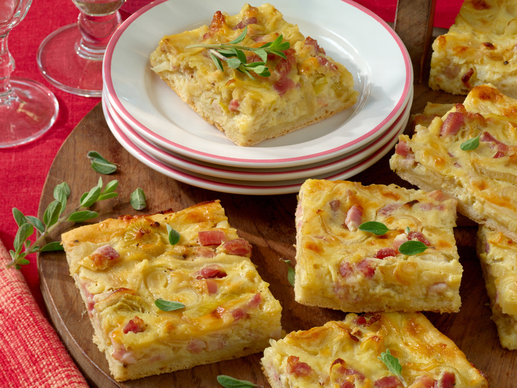 Mehrere Stücken vom Zwiebelkuchen auf einem Holzbrett mit einem kleinen weißen Teller und einem weiteren Stück davon darauf. Alles von schräg oben fotografiert.