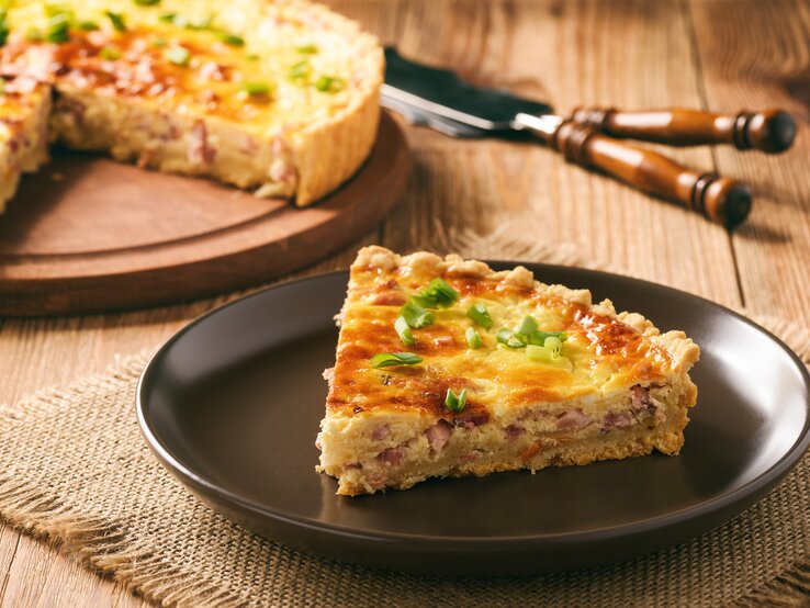  Ein Stück Quiche Lorraine auf einem dunklen Teller, garniert mit frischen Frühlingszwiebeln.