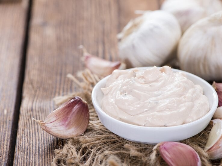 Schale mit Aioli auf Holztisch, daneben Knoblauchzehen. 
