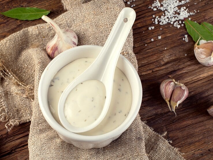 Weiße Schale mit einer cremigen Aioli mit Kräutern auf einem Holzbrett.